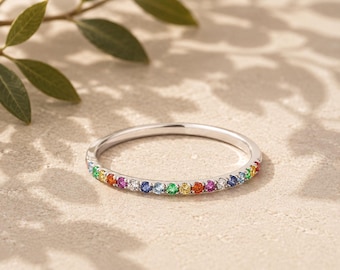 May include: A thin silver-toned metal band ring featuring a row of small, round, multi-colored gemstones in a rainbow sequence, including red, orange, yellow, green, blue, and purple stones set along the front half of the circular band.