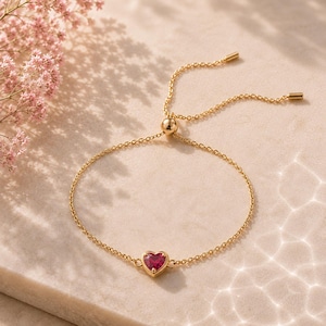 Puede incluir: Una pulsera de cadena en tono dorado con una piedra preciosa roja en forma de corazón en el centro. La pulsera incluye un mecanismo de cierre deslizante ajustable con dos cadenas finas que salen de una cuenta central para el ajuste.