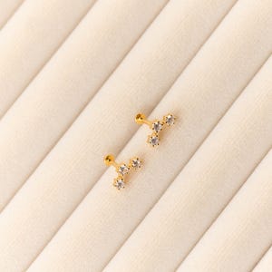 May include: Two gold-colored stud earrings, each featuring a cluster of three clear, sparkling gemstones. The earrings are displayed on a cream-colored, textured surface, creating a simple and elegant presentation. These earrings are a delicate jewelry item.