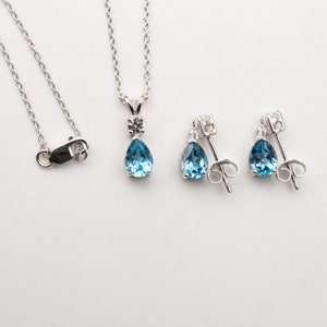 May include: A silver necklace and matching earrings set. The necklace has a teardrop-shaped, light blue gemstone pendant with a small, round accent stone. The earrings have a similar design. The jewellery is displayed on a white background.