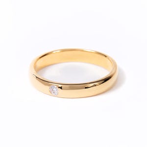 May include: A gold-colored ring with a single, small, clear gemstone. The ring has a smooth, polished surface and a classic band design. The gemstone is set into the band. The ring is displayed on a white background.