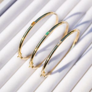 May include: Three gold bangle bracelets with different coloured gemstones. The top bracelet has an orange gemstone, the middle bracelet has a green gemstone, and the bottom bracelet has a clear gemstone.