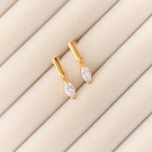 May include: A pair of gold-colored earrings. Each earring features a rectangular top with a dangling, marquise-cut, clear gemstone. The earrings are displayed on a cream-colored, textured surface.