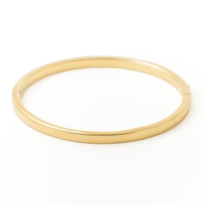 Könnte beinhalten: Ein goldfarbenes Armreif-Armband. Das Armband ist ein einfaches, glattes Band mit einem Scharnierverschluss. Der Schmuck ist vor einem weißen Hintergrund platziert.