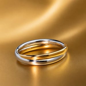 May include: A two-tone ring featuring a polished silver-colored band intertwined with a smooth gold-colored band. The interior of the silver band is clearly engraved with the text S925, indicating the metal composition of the jewelry piece.