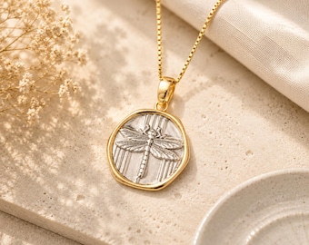 May include: A gold-toned necklace featuring a round, irregular pendant with a silver-toned dragonfly embossed in the center. The pendant is suspended from a delicate gold-toned box chain, creating a two-tone metallic appearance with a textured finish.