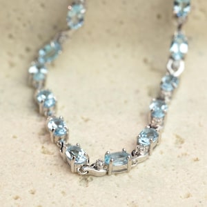 Könnte beinhalten: Ein silbernes Armband mit ovalen, hellblauen Edelsteinen. Die Edelsteine sind in einem Ketten-Design gefasst, mit kleinen Akzentsteinen zwischen jedem Oval. Das Armband wird auf einer neutralen Oberfläche präsentiert.