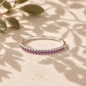 May include: A thin silver-toned band ring featuring a continuous row of small, round, vibrant magenta-pink gemstones set along the front half of the band. The metal has a polished finish, and the stones are evenly spaced in a shared prong setting.