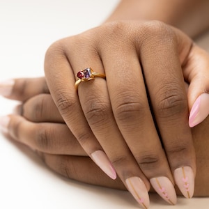 May include: A gold ring featuring a heart-shaped red gemstone and a square-cut purple gemstone. The ring is set on a gold band. The image shows the ring on a person's finger.
