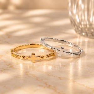 Puede incluir: Dos anillos finos de metal, uno dorado y otro plateado, cada uno con un pequeño dije de cruz integrado en la banda. Las bandas tienen un diseño texturizado con cuentas y pequeños segmentos rectangulares espaciados uniformemente.