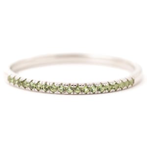 May include: A silver ring with a band of small, light green gemstones. The ring is set against a white background, highlighting the gemstones' color and the ring's design. The ring is a piece of jewelry.