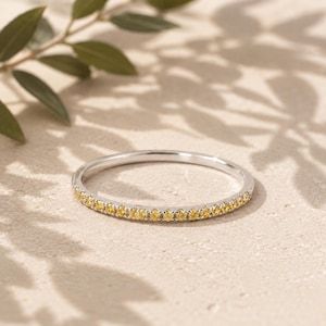 May include: A thin, polished silver-tone metal band ring featuring a single row of small, round, bright yellow gemstones set along the front half of the band. The ring has a smooth, reflective finish and a delicate, slender profile.