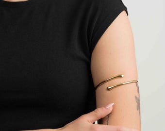 May include: A gold-toned metal arm cuff featuring a sleek, open-ended design with rounded, tapered tips. The polished, smooth band wraps partially around the upper arm, creating a minimalist, continuous curve that highlights a metallic finish.