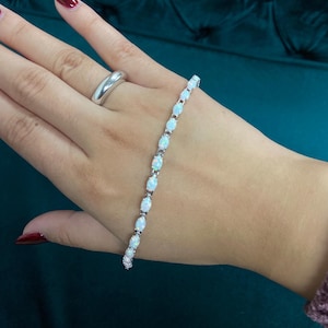 Puede incluir: Una pulsera de plata con ópalos ovalados engastados en un diseño delicado. La pulsera se muestra en una mano, destacando las piedras blancas e iridiscentes brillantes. Un anillo de plata también es visible en la mano, complementando el elegante estilo de la pulsera.