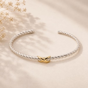 May include: A silver-toned cuff bracelet featuring a twisted rope-style band and a central gold-toned X-shaped accent. The bracelet has an open design with rounded ends, showcasing a polished metallic finish throughout the entire piece.