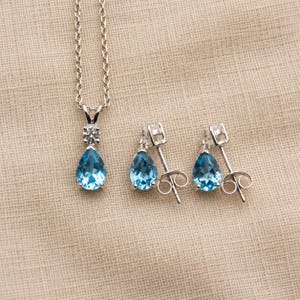 May include: A silver necklace and matching stud earrings set featuring pear-shaped blue gemstones. The necklace pendant includes a small round clear stone above the blue teardrop, while each earring displays a single pear-shaped blue gemstone.