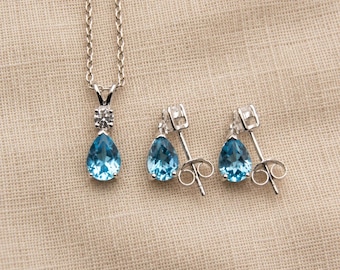 Puede incluir: Un conjunto de collar y pendientes de plata con gemas azules en forma de pera. El colgante del collar incluye una pequeña piedra clara redonda sobre la gota azul, mientras que cada pendiente muestra una única gema azul en forma de pera.
