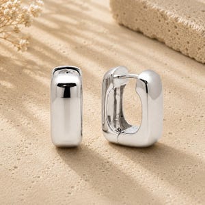 May include: A pair of polished silver-tone hoop earrings with a rectangular, chunky silhouette. The earrings feature a smooth, reflective metallic surface and a hinged post mechanism, showcasing a bold, geometric design with rounded corners.