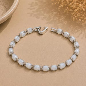 Puede incluir: Una pulsera de eslabones en tono plateado que presenta una serie de piedras cabujón blancas e iridiscentes de forma ovalada engastadas en marcos de metal. La pulsera incluye un cierre metálico seguro y está dispuesta en círculo.