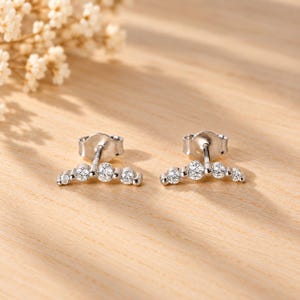 May include: A pair of silver-toned stud earrings, each featuring a curved bar design set with four round, clear, faceted stones of varying sizes. The earrings have a polished metallic finish and include standard post-style backings.