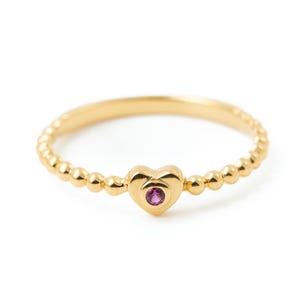 May include: A gold-colored ring with a heart-shaped center stone. The ring features a beaded band and a heart-shaped setting with a small, round, pink gemstone. The ring is set against a white background.