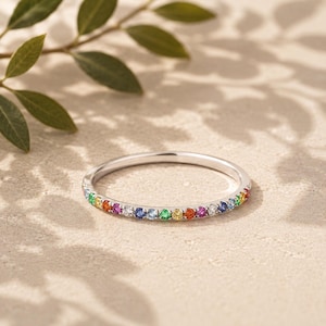 Op de afbeelding: Een dunne zilverkleurige metalen ring met een rij kleine, ronde, veelkleurige edelstenen in een regenboogvolgorde, waaronder rode, oranje, gele, groene, blauwe en paarse stenen die langs de voorste helft van de ring zijn gezet.