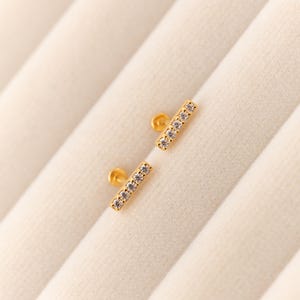 May include: Two gold-colored bar earrings, each adorned with a row of small, clear gemstones. The earrings have a post and backing. The background is a soft, off-white fabric.