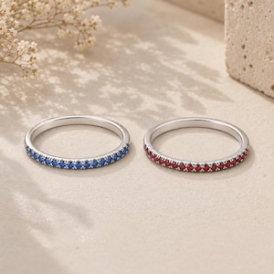 May include: Two thin metal rings featuring a pavé setting. One ring is adorned with a continuous row of small, round, vibrant blue stones, while the other ring features a matching row of small, round, deep red stones along the outer band.
