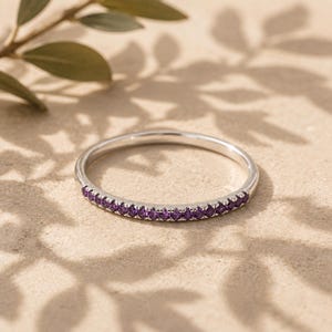 May include: A thin, polished silver-tone metal band ring featuring a row of small, round, vibrant purple gemstones set along the front half of the band. The ring has a smooth, reflective finish and a delicate, slender profile with a prong setting.