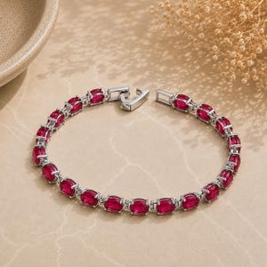 Könnte beinhalten: Ein silberfarbenes Tennisarmband mit einer Reihe von oval geschliffenen, leuchtend roten Edelsteinen, die miteinander verbunden sind. Das Armband verfügt über einen sicheren Metallverschluss und ist kreisförmig angeordnet.