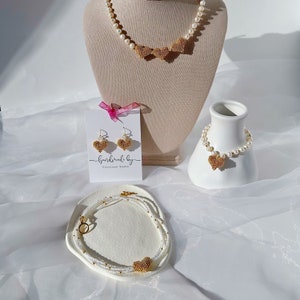 Puede incluir: Un conjunto de joyas de perlas con detalles de corazón de oro. El conjunto incluye un collar, una pulsera y pendientes. Las joyas se muestran sobre un fondo blanco.