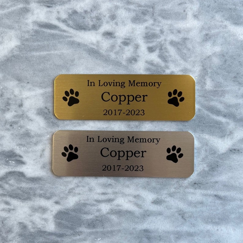 Op de afbeelding: Twee gouden en zilveren gedenkplaten met poot afdrukken en de tekst "In Loving Memory Copper 2017-2023".