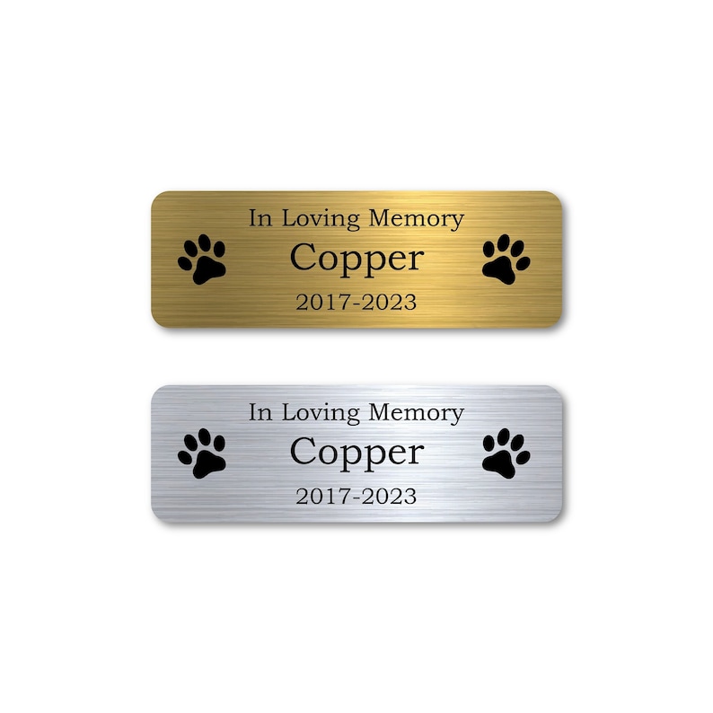 Op de afbeelding: Twee rechthoekige gedenkplaatjes voor huisdieren, een gouden en een zilveren, met de tekst "In Loving Memory Copper 2017-2023" en twee poot afdrukken.