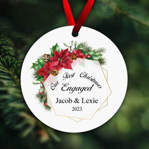 May include: White round Christmas bauble featuring a red poinsettia, green holly, and pine branches. The bauble has a gold geometric frame with the text "Our First Christmas Engaged Jacob & Lexie 2023".