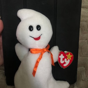 Può includere: Un peluche bianco a forma di fantasma beanie baby con una bocca rossa, occhi neri e un nastro arancione intorno al collo. Il beanie baby ha un'etichetta Ty rossa con le parole "Beanie Baby" e una stella.