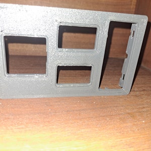 Puede incluir: Un panel de plástico gris con tres aberturas rectangulares y una sección lateral con pequeñas pestañas. El panel es probablemente un componente interior de un vehículo, posiblemente para interruptores o controles. El artículo está hecho de plástico gris mate.