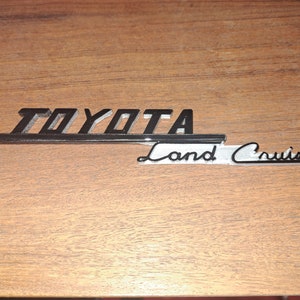 Puede incluir: Un emblema vintage de Toyota Land Cruiser, con el nombre de la marca en negrita y en negro sobre la escritura cursiva "Land Cruiser". El emblema tiene un acento plateado y est&aacute; colocado sobre una superficie de madera.