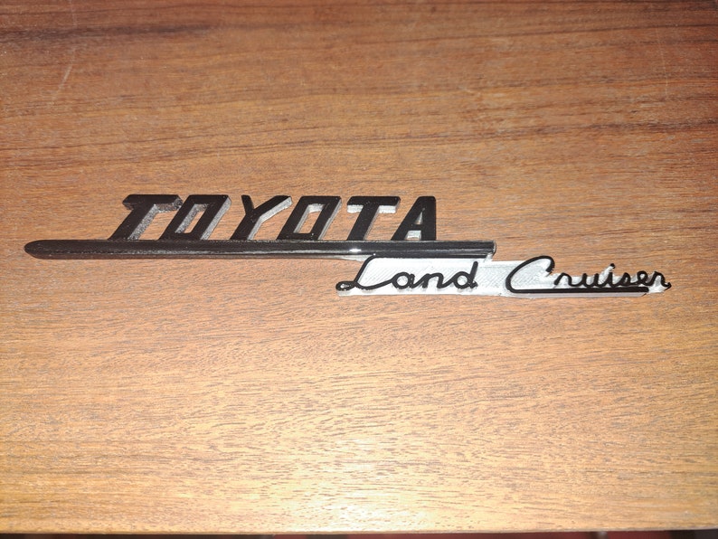 Puede incluir: Un emblema vintage de Toyota Land Cruiser, con el nombre de la marca en negrita y en negro sobre la escritura cursiva "Land Cruiser". El emblema tiene un acento plateado y est&aacute; colocado sobre una superficie de madera.