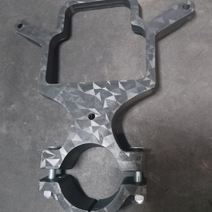May include: A dark gray carbon fiber mounting bracket featuring a geometric triangular pattern. The component includes a rectangular upper frame with two side arms and a circular lower clamp secured by two silver metal bolts and nuts.