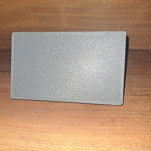 May include: A rectangular, grey object with a textured surface. The object is resting on a wooden surface. The item appears to be a small, solid block.