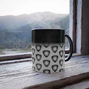 May include: A black and white heat-sensitive mug with a repeating rotary engine symbol pattern. The mug is on a weathered wooden windowsill, with a mountain view in the background. Steam rises from the mug.