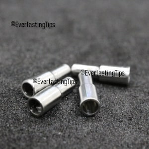 May include: Four silver metal arrowheads, also known as arrow points, are scattered on a dark gray surface.  The arrowheads are cylindrical with a pointed tip and a hollow base.