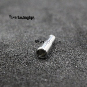 May include: A silver metal wire connector, also known as a wire crimp, is shown on a gray textured surface. The connector is cylindrical in shape with a small hole at one end.