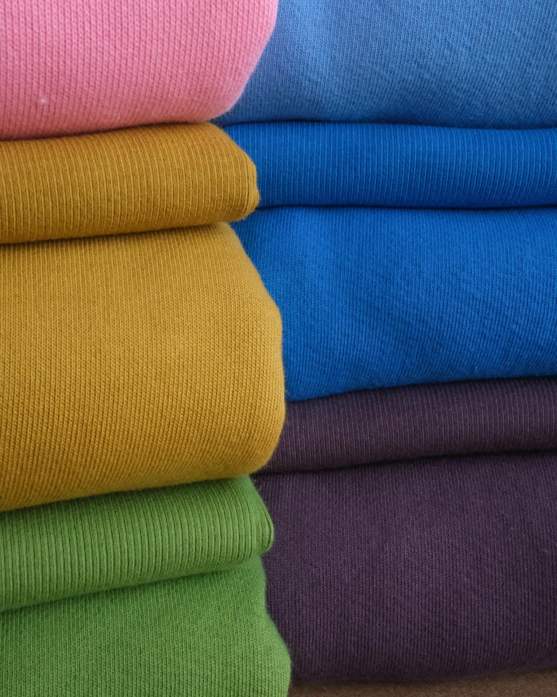 K&ouml;nnte beinhalten: Nahaufnahme von gefaltetem Stoff in verschiedenen Farben. Die Farben umfassen Rosa, Blau, Gold, Gr&uuml;n und Lila. Der Stoff scheint ein Strickmaterial zu sein, m&ouml;glicherweise Baumwolle oder eine Baumwollmischung. Die Falten erzeugen einen geschichteten Effekt, der die verschiedenen Farbt&ouml;ne zeigt.