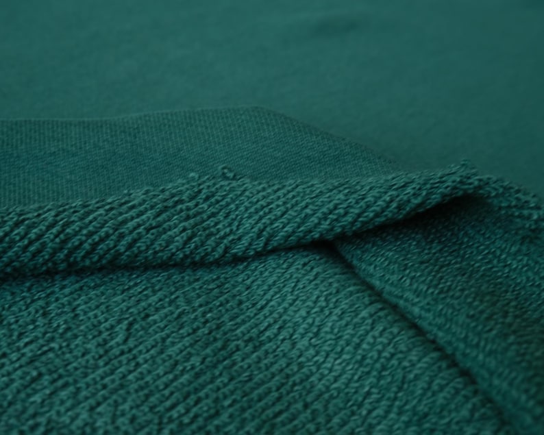 K&ouml;nnte beinhalten: Nahaufnahme eines teal-gr&uuml;nen Stoffes. Der Stoff hat eine strukturierte, geschlungene Oberfl&auml;che und einen gerippten Rand. Das Bild hebt die Textur und Farbe des Materials hervor, was auf ein Textil oder Kleidungsst&uuml;ck hindeutet.
