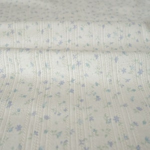 May include: Close-up of a cream-colored fabric with a ribbed texture. Tiny blue and green floral patterns are scattered across the surface. The fabric is slightly wrinkled, suggesting it is soft and pliable, suitable for clothing or home decor.