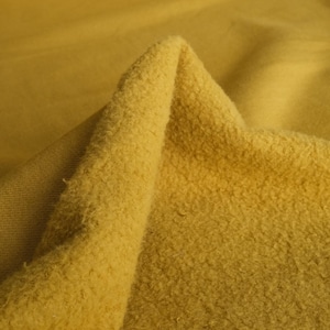 Pode incluir: Close-up de um tecido amarelo mostarda dobrado. O tecido tem uma superf&iacute;cie texturizada e difusa de um lado e uma textura lisa e tecida do outro. A ilumina&ccedil;&atilde;o destaca as diferentes texturas e tons de amarelo.