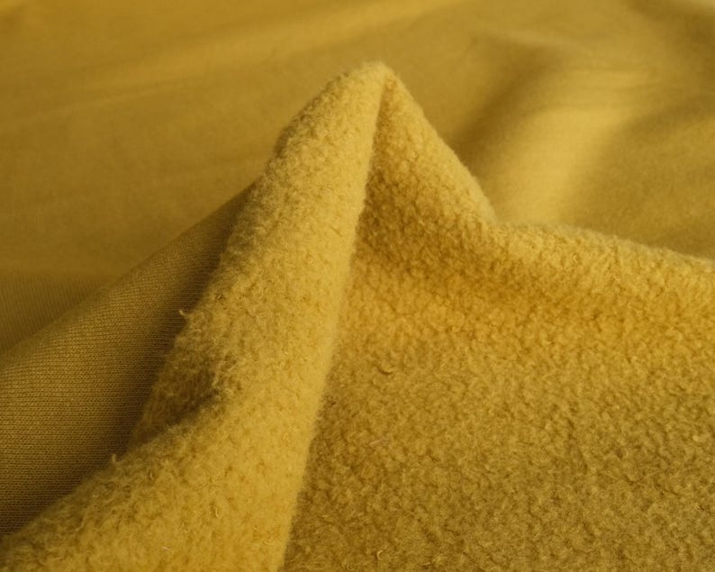 Pode incluir: Close-up de um tecido amarelo mostarda dobrado. O tecido tem uma superf&iacute;cie texturizada e difusa de um lado e uma textura lisa e tecida do outro. A ilumina&ccedil;&atilde;o destaca as diferentes texturas e tons de amarelo.