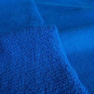 K&ouml;nnte beinhalten: Nahaufnahme von zwei blauen Stoffst&uuml;cken. Ein St&uuml;ck hat eine strukturierte, geschlungene Frottee-Optik, w&auml;hrend das andere glatt ist. Die Farbe ist ein leuchtendes, sattes Blau, wobei die Texturen Schatten und Tiefe erzeugen.