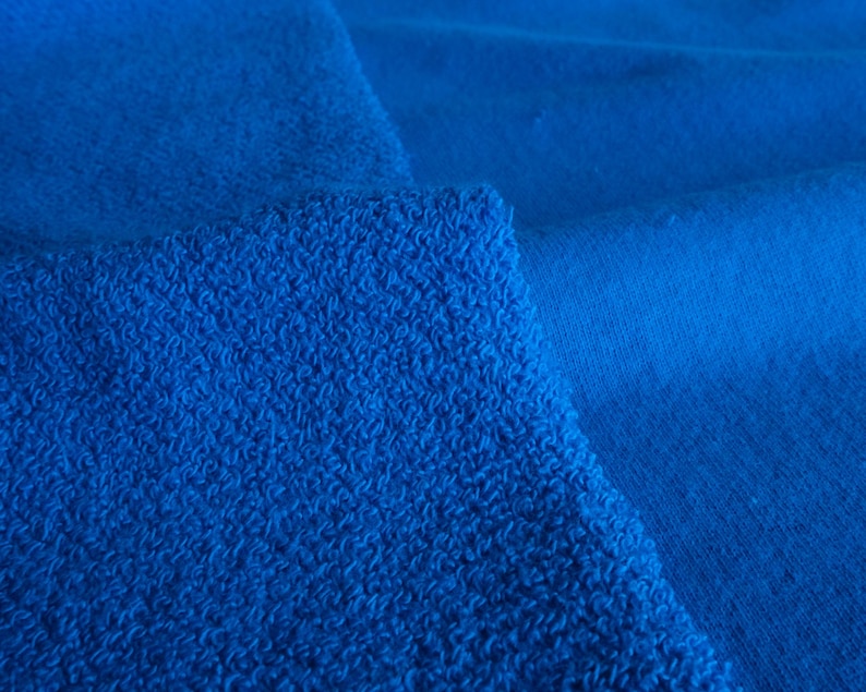 K&ouml;nnte beinhalten: Nahaufnahme von zwei blauen Stoffst&uuml;cken. Ein St&uuml;ck hat eine strukturierte, geschlungene Frottee-Optik, w&auml;hrend das andere glatt ist. Die Farbe ist ein leuchtendes, sattes Blau, wobei die Texturen Schatten und Tiefe erzeugen.